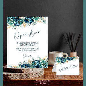 以下が含まれることがあります： 「Open Bar」の文字と、ティール色の花のアクセントが入った白い看板。看板には「私の特別な日を共有してくれてありがとう」と「今夜は私がお酒をご馳走します」というテキストも含まれています。また、「gluten free」と書かれた小さな看板もあります。