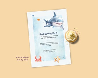 May include: A party invitation featuring a watercolour shark illustration. The invitation reads "Shark Sighting Alert!" and includes party details. A cupcake with white icing and sprinkles is next to the invitation.