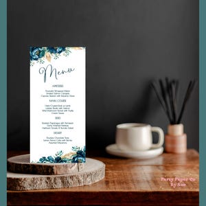 Può includere: Un biglietto del menu verticale con un design floreale blu e oro, con la parola "Menu". Il menu elenca antipasti, piatti principali, contorni e dessert. Il biglietto è esposto su una fetta di legno.