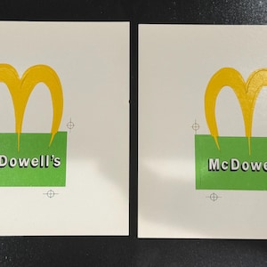 Può includere: Due biglietti quadrati bianchi con il logo McDowell's. Il logo presenta una 'M' gialla stilizzata sopra un rettangolo verde con la scritta 'McDowell's' in bianco. I biglietti hanno piccoli segni di registro negli angoli.