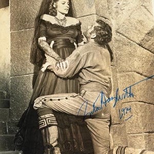 May include: A monochrome photograph featuring a man kneeling before a woman. The woman is in a dark dress with lace sleeves and a veil. The man wears a light-coloured shirt and trousers. The image is signed by Rita Hayworth and dated 1974.