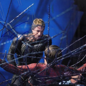 May include: A scene from a movie featuring two costumed characters, one in a black suit with spiderweb detailing and the other in a red and black Spider-Man suit, entangled in a web. The background is a blurred blue.