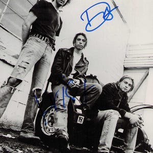Può includere: Una fotografia in bianco e nero della rock band Nirvana con tre membri. L'immagine include firme a inchiostro blu sulla stampa. I membri della band indossano abiti casual, tra cui jeans, magliette e una giacca di pelle.