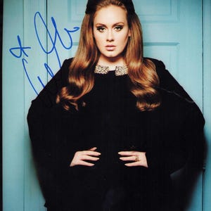 Puede incluir: Fotografía firmada de una mujer con cabello castaño rojizo ondulado y un vestido negro con cuello joya. El fondo es una puerta azul claro. La firma es en tinta azul.