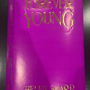 Könnte beinhalten: Ein lila Hardcover-Buch mit dem Titel "FOREVER YOUNG" in goldener Schrift. Der Name des Autors, Helen Sharp, ist ebenfalls in Gold gehalten. Der Buchrücken zeigt den Titel vertikal. Das Cover ist ein kräftiges, leuchtendes Lila, das einen auffälligen Kontrast zum goldenen Text bildet.