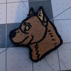 Op de afbeelding: Een geborduurde patch met een cartoon hond, bruin met zwarte contouren. De hond kijkt naar links en heeft een vrolijke uitdrukking.