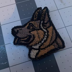 May include: A brown and black embroidered patch of a German Shepherd dog's head. The dog is looking to the left and has a white collar.
