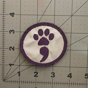 以下が含まれることがあります： 紫と白の刺繍パッチで、足跡とセミコロンが描かれています。パッチは円形で、白い背景に紫色の縁取りがあります。