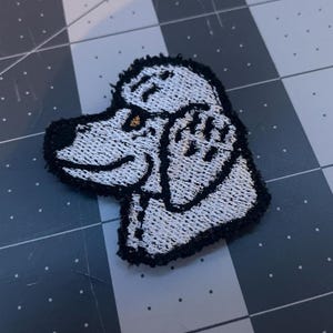 May include: White embroidered patch of a poodle with black outline. The poodle's head is tilted to the side and its eyes are closed.