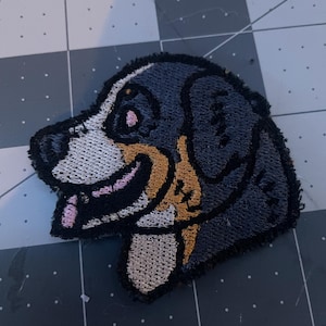 Puede incluir: Parche bordado de la cabeza de un perro de montaña bernés, con pelo negro, blanco y marrón. El perro está sonriendo con la lengua fuera.