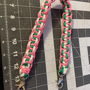 Puede incluir: Una correa trenzada en rosa, blanco y verde, con cierres giratorios plateados en cada extremo. La correa está hecha de cordones entrelazados, creando un patrón texturizado. El fondo es una esterilla de corte negra y gris.