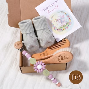 May include: A baby gift set in a cardboard box. The set includes gray star-shaped baby socks, a wooden comb and brush, and a pacifier clip with a flower design. A card with a unicorn illustration reads "Welcome to the world."