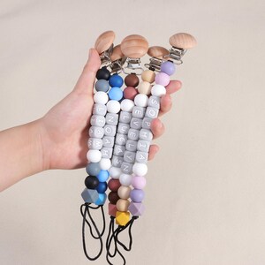 May include: A set of five silicone baby teethers with wooden clips. Each teether is a string of colorful beads with a gray cube bead that has a name on it. The names are Sophia, Evelyn, Lucas, Noah, and Liam.