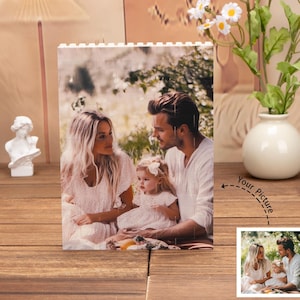 May include: A white brick-style photo frame with a picture of a family of three in a garden setting. The frame is on a wooden surface with a vase of white flowers in the background. The text "Your Picture" is written on the image.