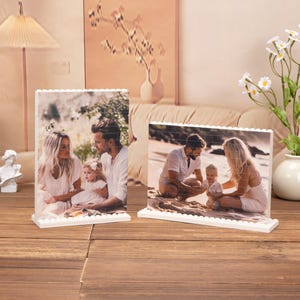 Op de afbeelding: Twee witte fotolijsten met een witte achtergrond en een gezin van drie op een strand. Het gezin bestaat uit een man, een vrouw en een kind. De man en de vrouw zitten op het zand, en het kind staat tussen hen in. De man draagt een blauw shirt en een korte broek, en de vrouw draagt een witte jurk. Het kind draagt een blauw shirt en een korte broek.