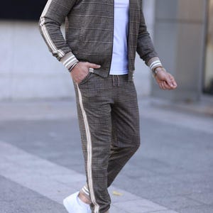 May include: A brown and white plaid tracksuit with white stripes down the sides of the pants. The jacket has a zipper closure and a ribbed collar. The model is wearing white sneakers.