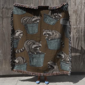 May include: A woven throw blanket with a brown background and a repeating pattern of raccoons in blue buckets.