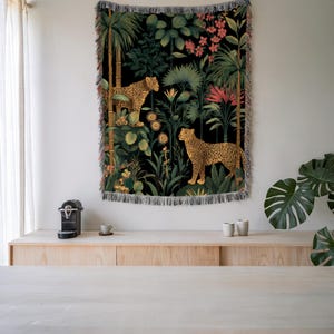 May include: A woven tapestry featuring a jungle scene with two leopards amidst lush greenery and flowers. The background is black, and the tapestry has a fringed edge. The leopards have a spotted pattern, and the plants are in shades of green, yellow, and pink.
