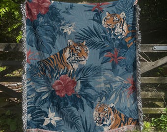 May include: A woven throw blanket featuring a repeating pattern of tigers, large tropical leaves, and red hibiscus flowers on a dark blue background. The blanket has a textured finish and decorative fringe along the top and bottom edges.
