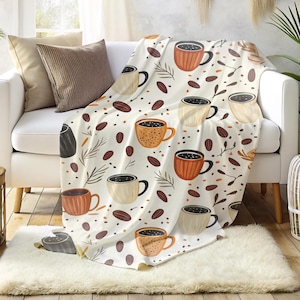 Könnte beinhalten: Eine weiße Decke mit Kaffeemotiv. Die Decke zeigt ein sich wiederholendes Muster aus Kaffeetassen in Braun- und Beigetönen, Kaffeebohnen und Zweigen. Die Decke ist über ein weißes Sofa drapiert.