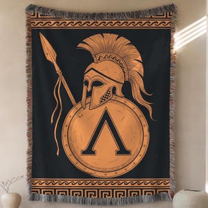 May include: A woven throw blanket featuring a black background with a large orange Spartan helmet, a spear, and a circular shield displaying the Greek letter Lambda. The top and bottom borders have orange wave and geometric meander patterns.