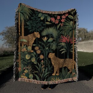 May include: A black woven throw blanket featuring a jungle scene with two leopards, palm trees, and tropical flowers. The blanket has a fringed edge and is made of a soft, woven material. The color palette includes shades of green, brown, and pink.