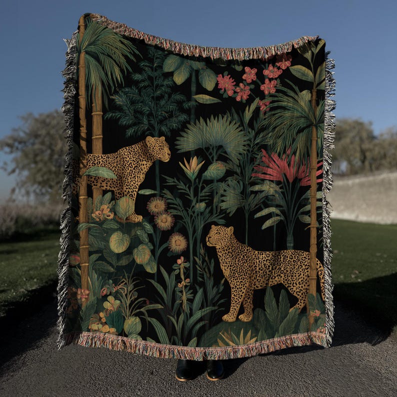 May include: A black woven throw blanket featuring a jungle scene with two leopards, palm trees, and tropical flowers. The blanket has a fringed edge and is made of a soft, woven material. The color palette includes shades of green, brown, and pink.