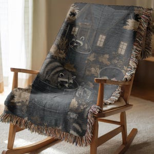 May include: A woven throw blanket featuring a dark, nocturnal scene of two raccoons foraging in metal trash cans outside a house. The design includes scattered autumn leaves in shades of brown and tan, with fringed edges on all sides.