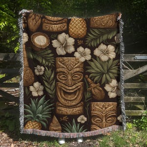 Peut inclure: Une couverture tissée marron avec un motif répété de masques tiki, d'ananas, de fleurs d'hibiscus et de feuillage tropical. La couverture a une bordure à franges et est exposée à l'extérieur.