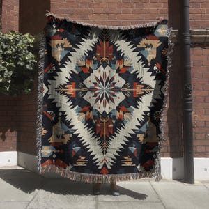 May include: A woven throw blanket with a geometric pattern in shades of black, white, red, and blue. The pattern features a central starburst design with repeating triangles and diamond shapes.
