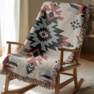 Pu&ograve; includere: Una coperta tessuta con un motivo geometrico nei toni del nero, rosso spento e azzurro su fondo crema. La coperta presenta una superficie testurizzata e frange decorative lungo i bordi, drappeggiata su una sedia a dondolo in legno.