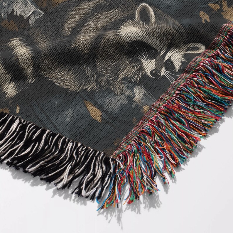 May include: A woven throw blanket featuring a detailed graphic of a raccoon against a dark background. The blanket has a textured appearance with a prominent fringe border along the edges, showcasing a mix of multicolored threads in the fringe.