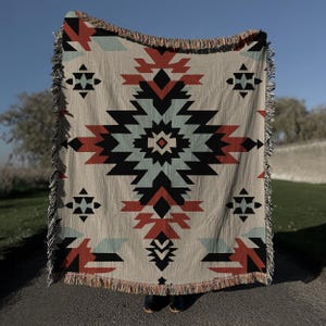 Pu&ograve; includere: Una coperta tessuta caratterizzata da un grande motivo geometrico a rombi centrato, nei toni del terracotta, azzurro e nero su sfondo beige. La coperta presenta un bordo sfrangiato sulla parte superiore e inferiore, con motivi geometrici.