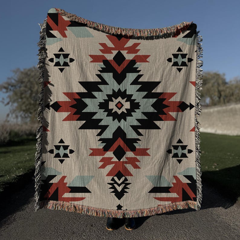 Pu&ograve; includere: Una coperta tessuta caratterizzata da un grande motivo geometrico a rombi centrato, nei toni del terracotta, azzurro e nero su sfondo beige. La coperta presenta un bordo sfrangiato sulla parte superiore e inferiore, con motivi geometrici.