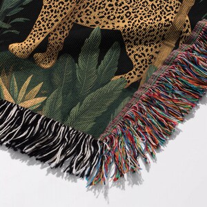 May include: Woven throw blanket featuring a leopard design with a black background and green foliage. The blanket has a fringed edge with a mix of colorful threads. The leopard's spots are a mix of tan and brown.