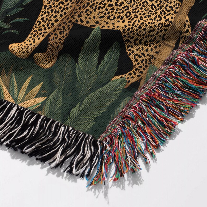 May include: Woven throw blanket featuring a leopard design with a black background and green foliage. The blanket has a fringed edge with a mix of colorful threads. The leopard's spots are a mix of tan and brown.
