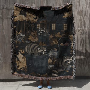 May include: A woven throw blanket featuring a dark forest scene with multiple raccoons foraging near trash cans and a cabin. The design includes golden-brown autumn leaves, dark grey barrels, and a fringed edge on all sides of the rectangular textile.