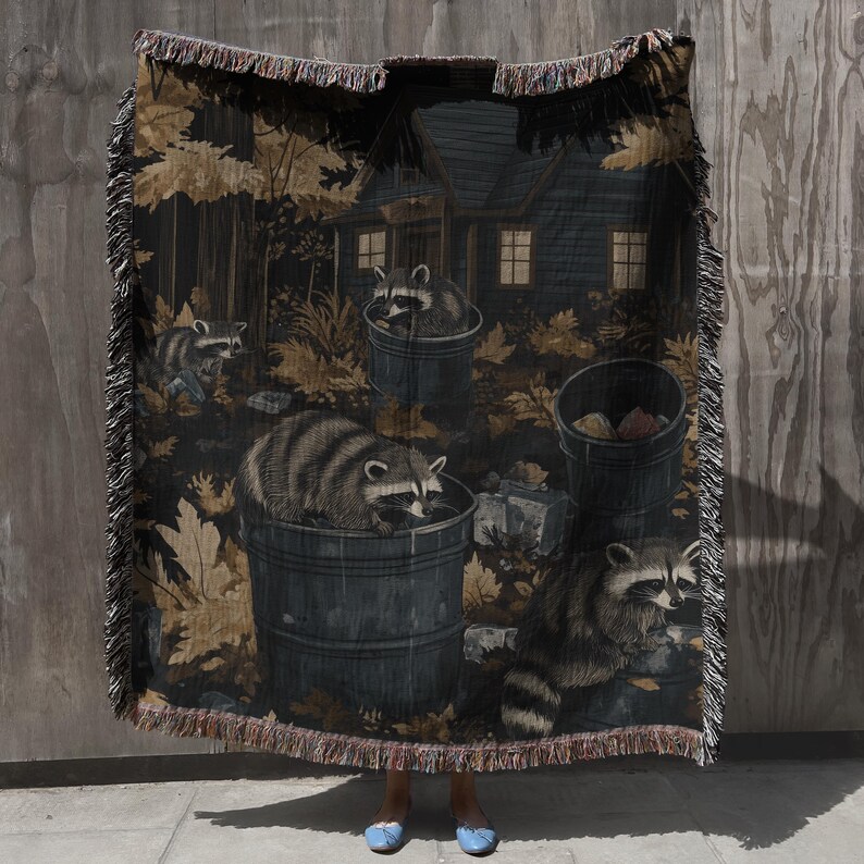 May include: A woven throw blanket featuring a dark forest scene with multiple raccoons foraging near trash cans and a cabin. The design includes golden-brown autumn leaves, dark grey barrels, and a fringed edge on all sides of the rectangular textile.