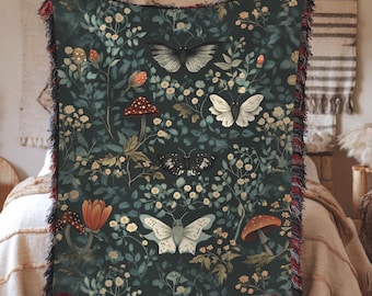 May include: A woven throw blanket featuring a dark green background with a botanical pattern of white flowers, green leaves, red and brown mushrooms, and four butterflies in shades of black, white, and cream, finished with a multi-colored fringed edge.