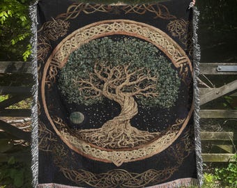 May include: A black woven throw blanket featuring a central circular design of a gnarled tree with green foliage and exposed roots. The tree is enclosed in a tan Celtic knotwork border, with additional intricate knotwork patterns in the corners.