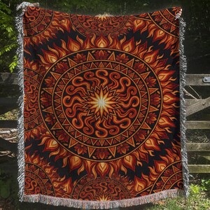 May include: A rectangular woven throw blanket featuring a large central circular mandala design in shades of orange, red, and yellow against a black background. The pattern includes radiating flame-like motifs and white fringed edges on all sides.