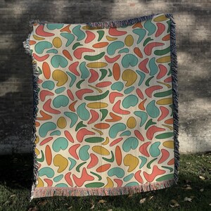 May include: A woven throw blanket featuring a repeating pattern of abstract organic shapes in shades of teal, coral, mustard yellow, and forest green on a cream background, finished with decorative fringe along the top and bottom edges.
