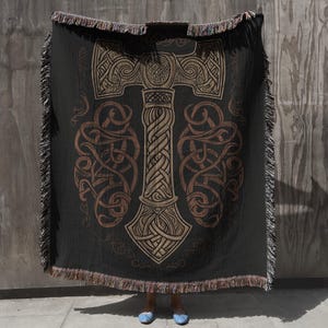 May include: Black throw blanket featuring a large, detailed illustration of Thor's hammer, Mjolnir, in shades of brown and tan. The design includes intricate knotwork and Celtic patterns. The blanket has a fringed edge.