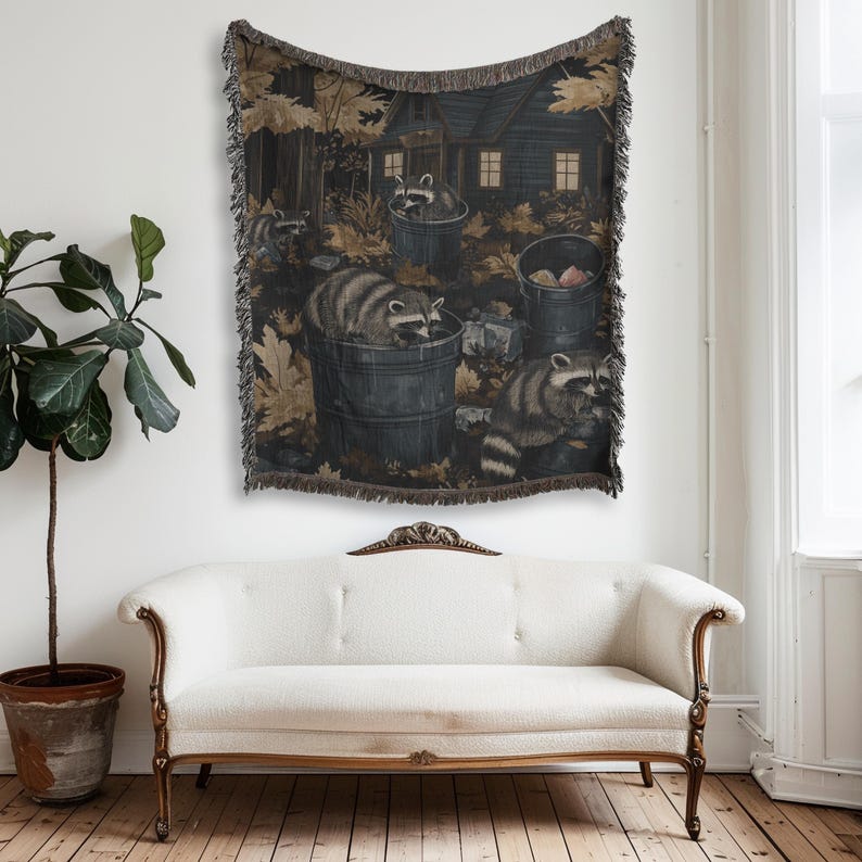 May include: A woven throw blanket featuring a dark, nocturnal forest scene with multiple raccoons foraging in and around metal trash cans. The design includes a rustic cabin in the background, fallen autumn leaves, and a decorative fringed edge.