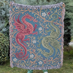 May include: A woven throw blanket featuring a teal background with a repeating pattern of a red dragon and a green dragon intertwined with gold scrolling vines and white flowers. The blanket has a decorative fringed edge on the top and bottom.