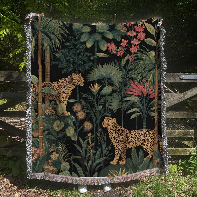 May include: A black throw blanket featuring a jungle scene with two leopards, palm trees, and tropical flowers. The blanket has a fringed edge and is made of a soft, woven material. The color palette includes shades of green, brown, and pink.