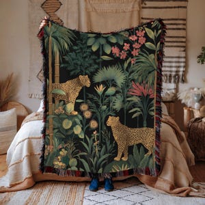 May include: A black woven throw blanket featuring a jungle scene with two leopards, lush green foliage, and pink flowers. The blanket has a fringed edge with red and black accents. The design is detailed and vibrant, creating a tropical aesthetic.