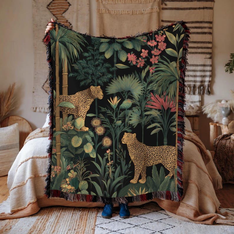 May include: A black woven throw blanket featuring a jungle scene with two leopards, lush green foliage, and pink flowers. The blanket has a fringed edge with red and black accents. The design is detailed and vibrant, creating a tropical aesthetic.