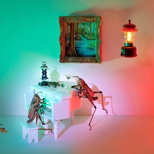 May include: Two brown and green grasshoppers playing a white piano with a glass of liquid on the piano. A framed painting of a swamp scene hangs on the wall behind the piano. A small, red lantern is on the wall to the right of the piano.