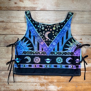 May include: A black sleeveless crop top featuring a vibrant celestial print with moons, suns, and planets in shades of blue, purple, and teal. The garment has a scoop neckline and is secured on both sides with thin black fabric ties in a lace-up style.